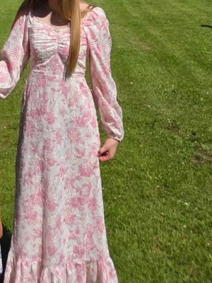 Lång blommig klänning med puffärm -  långklänning i vitt och rosa med blommigt mönster. Klänningen har puffiga långärmar, markerad midja och volang längst ner. Söt omlottdetalj vid bysten och lätt rynkade detaljer. Nyskick