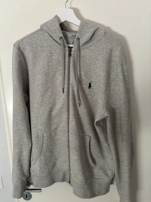 Grå hoodie från Polo Ralph Lauren - Snygg grå hoodie från Polo Ralph Lauren med dragkedja, huva med dragsko och två fickor framtill. Klassisk logga broderad på bröstet. Tillverkad i mjukt bomullsmaterial som känns skönt mot huden. Perfekt för en avslappnad och stilren look.
