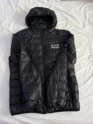Svart pufferjacka från EA7 Emporio Armani - Svart pufferjacka med huva från EA7 Emporio Armani. Jackan har quiltad design, dragkedja framtill och logga på bröstet. Tillverkad i glansigt, lätt material som håller värmen under kalla dagar. Perfekt för vintern och ger en sportig look.
