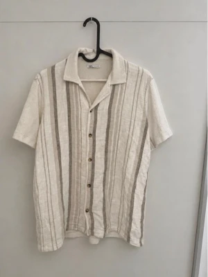 Randig linneskjorta från Zara - Säljer en snygg kortärmad skjorta från Zara i beige och vit med vertikala ränder. Skjortan har klassisk krage, knappar framtill och är gjord i luftigt linne – perfekt för varma dagar.