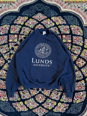 Mörkblå sweatshirt Lunds Universitet - Mörkblå sweatshirt med tryck från Lunds Universitet. Tröjan har rund hals, långa ärmar och ribbade muddar vid ärmslut och nederkant. Klassisk collegestil med stort vitt universitetsmärke på bröstet.
