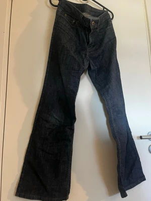 Mörkblå lågmidjade jeans - Säljer ett par mörkblå lågmidjade jeans. Jeansen har utsvängda ben och är tillverkade i denim med synliga sömmar, unik knapp och mönster på fickorna. Perfekta för dig som gillar en retroinspirerad look.