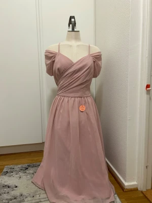 Ljusrosa långklänning med v-ringning - Romantisk ljusrosa långklänning med v-ringad omlottdesign och tunna axelband. Klänningen har offshoulder-detaljer och ett svepande, luftigt fall. Perfekt för speciella tillfällen där du vill känna dig extra fin. Ny med prislapp.