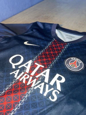 PSG matchtröja Nike blå/röd - Snygg PSG matchtröja från Nike i mörkblått med röd och vit rand i mitten. Tröjan har klubbmärke på bröstet, Qatar Airways-tryck och klassisk rund hals. Tillverkad i lätt och ventilerande material, perfekt för fotboll eller träning.