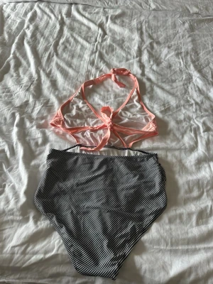 Randig bikini med hög midja - Säljer en bikini med ljusrosa omlottopp som knyts i nacken och ryggen, samt svarta och vita smalrandiga bikinitrosor med hög midja. Toppens omlottdesign ger en snygg och modern look. Perfekt för lata dagar på stranden!