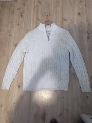 Vit kabelstickad tröja med halv dragkedja - Säljer en vit kabelstickad tröja med halv dragkedja i kragen. Tröjan har långa ärmar, ribbade muddar och nederkant samt ett klassiskt mönster. Perfekt för dig som gillar en clean och stilren look.
