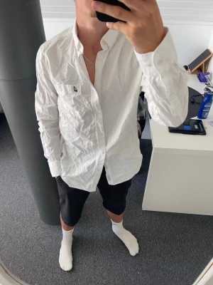 Vit skjorta peak performance  - Klassisk vit skjorta från Peak Performance med lång ärm och knappstängning framtill. Skjortan har en bröstficka med den ikoniska loggan broderad. Tillverkad i bomull och har en normal passform. Perfekt till många olika stilar.