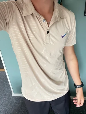 Beige pikétröja från Nike - träningspike golfpike  - Snygg beige pikétröja från Nike med klassisk krage och knappar framtill. Tröjan har korta ärmar och en diskret Nike-logga på bröstet. Tillverkad i ett lätt och bekvämt material som passar perfekt till träning eller sportiga outfits.