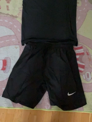 Svart träningsset från Nike - Säljer ett svart träningsset från Nike med t-shirt och shorts. Båda plaggen har den klassiska Nike-loggan i vitt. Tillverkade i ett lätt och snabbtorkande material som passar perfekt för träning eller sportaktiviteter.