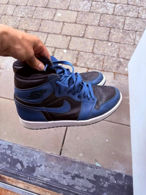 Nike Air Jordan 1 blå och svarta - Snygga Nike Air Jordan 1 sneakers i blått och svart läder med vita sulor. Klassisk high-top modell med ikonisk vinge-logga på sidan och matchande blå skosnören. Perfekta för dig som gillar streetwear och vill sticka ut.