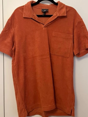 Orange frotté t-shirt H&M - Säljer en orange t-shirt från H&M i mjuk frotté med korta ärmar och en bröstficka. Skön och avslappnad passform med klassisk krage och slitsar i sidorna. Perfekt för en chill och trendig look.