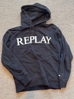 Svart hoodie från Replay med tryck - Snygg svart hoodie från Replay med stor vit logga på bröstet. Klassisk modell med huva och dragsnören, ribbade muddar vid ärmslut och nederkant. Pris kan diskuteras!!!