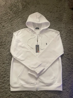 Vit hoodie från Polo Ralph Lauren - Snygg vit hoodie från Polo Ralph Lauren med dragkedja framtill och klassisk huva med dragsko. Ikoniska lilla loggan broderad på bröstet. Mjuk bomullskänsla och två fickor framtill. Perfekt för en clean och stilren look.