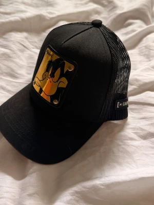 Svart keps Daffy Duck x Capster - Säljer en svart keps från Capster. Framtill sitter en gul metallplatta med Daffy Duck-motiv och texten 'DAFFY'. Broderad Daffy Duck-text på sidan. Justerbar snapback och böjd skärm. 