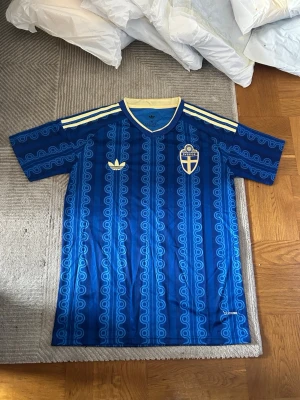 Blå Sverige fotbollströja Adidas - Säljer en blå fotbollströja från Adidas med Sveriges landslagsemblem på bröstet. Tröjan har retroinspirerat mönster, gula detaljer och klassiska Adidas-ränder på axlarna. Tillverkad i lätt och ventilerande material, perfekt för träning eller match.