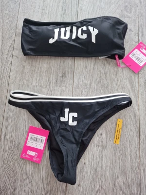 Svart bandeau bikini från Juicy Couture - Snygg svart bikini från Juicy Couture med bandeau-topp och vita tryckdetaljer. Toppen har texten JUICY och trosan har JC-logga framtill. Båda delar har vita ränder och är tillverkade i syntetmaterial. Perfekt för stranden eller poolen!
