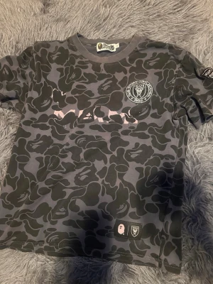 Camouflage t-shirt från A Bathing Ape - Säljer en grå och svart camouflage t-shirt från A Bathing Ape x Inter Miami. T-shirten har tryckt och broderad logga på bröstet samt patch på ärmen och nedtill. Klassisk rund hals och korta ärmar. Perfekt för dig som gillar streetwear. Eller fotboll 