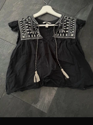 Svart boho-blus med broderi - Svart blus i boho-stil med vita broderier och tofsar framtill. Lös passform, korta ärmar och djup v-ringning. Dekorativa detaljer runt halsen och axlarna ger en unik look. Perfekt för en avslappnad och trendig stil.