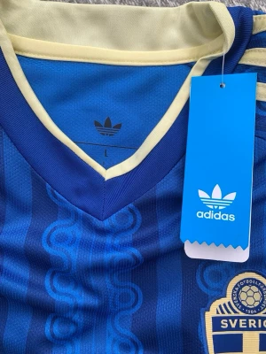 Blå Sverige tröja från Adidas - Säljer en blå fotbollströja från Adidas med Sveriges landslagsemblem på bröstet. Tröjan har gula detaljer, V-ringad krage och klassiska Adidas-ränder på axeln. Materialet är lätt och andas, perfekt för träning eller match.