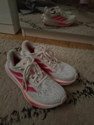 Vita med rosa detaljer träningsskor adidas  - Snygga vita Adidas träningsskor med klassiska tre rosa ränder på sidan och rosa detaljer på sulan. Jag har använt de ca 3 gånger på gymmet så de är i väldigt fint skick!! Kan fixa fler bilder bara att höra av sig. Kan även gå ner i pris vid snabb affär! Nypris ca 1100