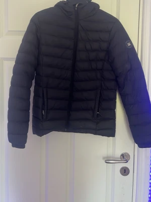 Svart dunjacka med huva - Snygg svart dunjacka med huva och dragkedja framtill. Jackan har två fickor med dragkedja och quiltad design. Märke syns på ärmen. Perfekt för kalla vinterdagar och riktigt skön att bära.