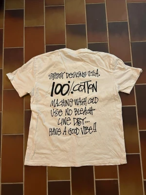 Vit Stüssy t-shirt med tryck - Vit t-shirt från Stüssy med svart logga på bröstet och stort grafiskt tryck på ryggen. Klassisk rund hals och korta ärmar. Tillverkad i 100% bomull för en skön och avslappnad känsla. Ni vet varför den är så billig… storlek enligt tag är M men sitter som XS-S