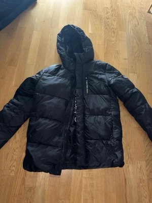 Svart pufferjacka från Calvin Klein - Snygg svart pufferjacka från Calvin Klein Jeans med huva och dragkedja framtill. Jackan har en logga på bröstet och ett CK-märke på ärmen. Tillverkad i glansigt material som ger en modern look och håller dig varm under kalla dagar. Köpt äkta från kidsbrandstore