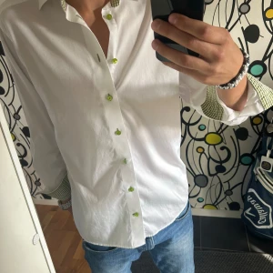 Vit skjorta från Eton - En elegant vit skjorta från Eton med gröna knappar och grönt rutigt mönster på insidan av kragen och manschetterna. Skjortan har långa ärmar och en modern passform. Storlek L, väldigt bra skick 