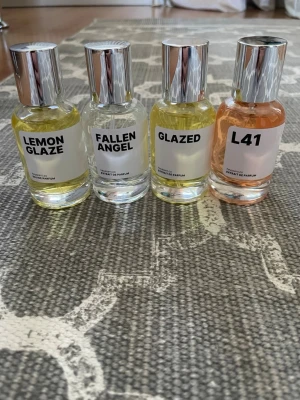 Fyra parfymer från Heyscent - Fyra flaskor parfym med moderna, minimalistiska etiketter och silverfärgade lock. Doftnamnen är LEMON GLAZE, FALLEN ANGEL, GLAZED och L41. Flaskorna är genomskinliga och innehållet varierar i färg från klart till gult och ljusrosa. Inspirerade av Lira, Bianco latte, Love dont be shy och Angels share