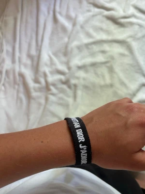 Svart Christian Dior tygarmband - Snyggt svart armband från Christian Dior med vit broderad text 'J'ADIOR'. Armbandet är gjort i tyg och har en enkel, stilren design som passar perfekt till din vardagsstil. Justerbar knytning för flexibel passform.