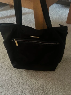 Svart väska från Victoria Secret - Säljer en snygg väska från Victoria Secret med gulddetaljer. Inuti är det leopardmönster samt ficka. Väskan har dragkedja.