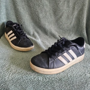 Svarta Adidas sneakers med vita ränder - Klassiska svarta Adidas sneakers med de ikoniska tre vita ränderna på sidorna och vit sula. Skorna har lågt skaft och snörning framtill. Ovandelen är i syntetmaterial och sulan är i gummi. Perfekta för en sportig och avslappnad stil.