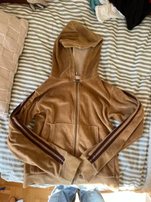 Brun velourhoodie med ränder - Snygg brun hoodie i mjuk velour med dragkedja framtill och två fickor. Huva med snörning och mörkbruna dubbla ränder längs ärmarna ger en retrovibe.