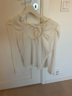 Vit långärmad topp med knyt - Snygg vit långärmad topp med rundad ringning och knyt fram. Toppen har lätt puffade axlar och är gjord i ett tunt, ribbat material som ger en skön och trendig look. Perfekt att styla med jeans eller kjol.