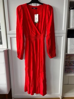 Röd omlottklänning från BIKBOK - Snygg röd långklänning från BIKBOK med omlottmodell och v-ringning. Klänningen har långa ärmar och markerad midja med smockdetalj. Perfekt för dig som vill sticka ut med en färgstark look.