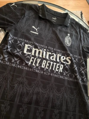  AC Milan tröja  - Snygg svart AC Milan matchtröja i samarbete med Pleasures och Puma. Tröjan har detaljerat grafiskt mönster, klubbmärke på bröstet och vit Emirates Fly Better-logga. Tillverkad i lätt och ventilerande material, perfekt för fotboll eller streetwear.