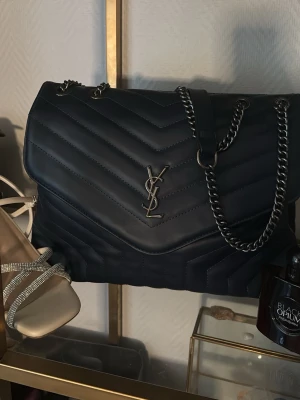 Mörkblå YSL handväska med silver kedja - Säljer en svart handväska från Yves Saint Laurent med quiltat mönster och silverfärgad kedjerem. Väskan har YSL-logga i metall framtill och en klassisk, stilren design. Perfekt för dig som gillar lyxiga accessoarer och tidlös stil.