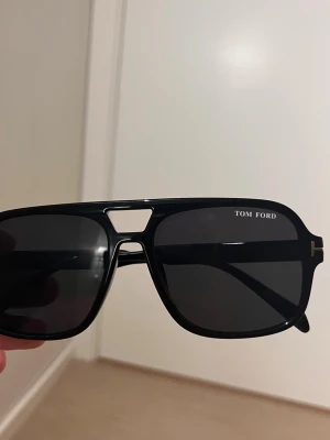 Svarta solglasögon från Tom Ford - Snygga svarta solglasögon från Tom Ford med klassisk pilotform och mörka glas. Ramen är i blank plast och har en cool detalj med metallinslag vid gångjärnen. Perfekta för att höja din stil på sommaren.