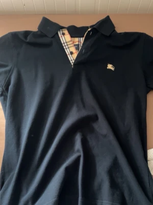 Svart pikétröja från Burberry - Svart pikétröja från Burberry med klassiskt rutigt tyg vid knäppningen och liten guldfärgad logga på bröstet. Skjortan har korta ärmar och är tillverkad i mjuk bomull. Perfekt för en stilren look.