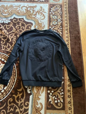 Svart sweatshirt från Dsquared2 - Cool svart sweatshirt från Dsquared2 med stort ton-i-ton broderat logomotiv på bröstet. Tröjan har rund hals, långa ärmar och ribbade muddar vid ärmslut och nederkant. Perfekt för en avslappnad och trendig look.