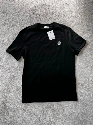 Svart Moncler t-shirt med logga - Svart t-shirt från Moncler med klassisk rund halsringning och korta ärmar. T-shirten har en broderad Moncler-logga på bröstet och är tillverkad i mjuk bomull. Perfekt för en stilren och enkel look. Ej äkta!!