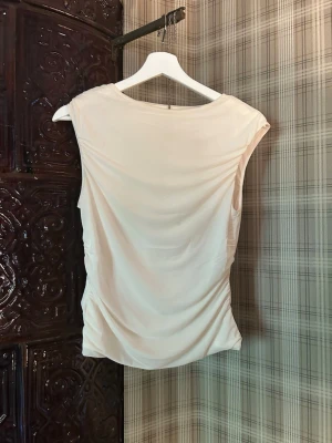 Beige ärmlös linnetopp - Säljer en stilren beige linnetopp med rund halsringning och draperad passform. Toppen är ärmlös och har en mjuk, lätt struktur som passar perfekt till många olika outfits.