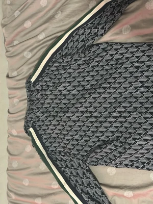 Mönstrad zip tröja  från Lacoste - Snygg zip tröja från Lacoste med svartvitt geometriskt mönster och dragkedja framtill. Tröjan har gröna och vita ränder längs ärmarna och klassisk Lacoste-logga på bröstet. Tillverkad i lätt material som passar perfekt till streetwear och sportig stil.