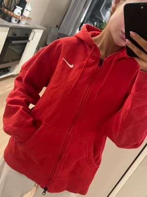 Röd zip hoodie från Nike - Röd hoodie från Nike med dragkedja framtill och vit broderad swoosh-logga på bröstet. Klassisk huva med dragsko och stora fickor framtill. Tillverkad i mjukt bomullsmaterial som är skönt att ha på sig. Är unisex så passar alla, knappt använd så jätte fint skick.
