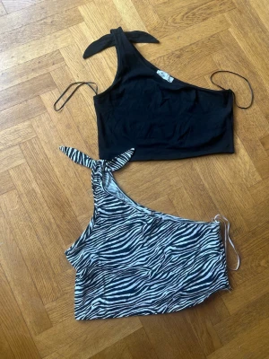 Oneshoulder toppar med knyt - Två trendiga oneshoulder toppar, en i svart och en med zebramönster. Båda har knyt vid axeln och är croppade. Perfekta för att skapa en cool och unik look. Materialet känns mjukt och stretchigt, vilket gör dem bekväma att bära.