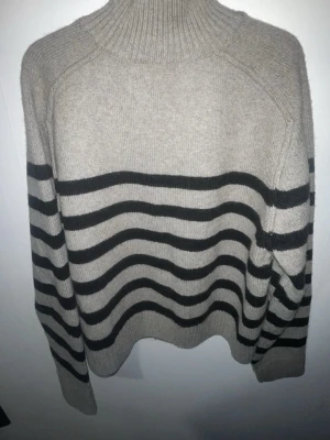 Beige stickad tröja med svarta ränder - Stickad tröja i beige med svarta horisontella ränder och hög ribbad krage. Långärmad och mjuk, perfekt för lager-på-lager. Tröjan har en avslappnad passform och ribbade muddar vid ärmslut och nederkant. Materialet ser ut att vara ull eller ullblandning.