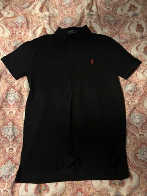 Svart pikétröja från Polo Ralph Lauren - Klassisk svart pikétröja från Polo Ralph Lauren med korta ärmar och röd broderad logga på bröstet. Tröjan har krage och knäppning med två knappar. Perfekt för en stilren look.