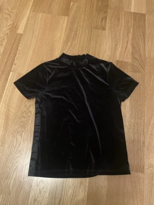 Svart velour t-shirt med hög krage - En svart t-shirt i mjuk velour med korta ärmar och en lite högre krage. T-shirten har en stilren och enkel design som ger en lyxig känsla. Perfekt för dig som gillar att sticka ut med materialvalet.