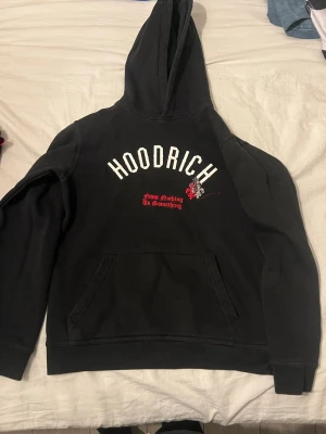 Svart hoodie från Hoodrich - Svart hoodie från Hoodrich med stor vit logga framtill och texten 'From Nothing To Something' i rött. På ryggen står det 'product of the streets' i vit gotisk stil. Klassisk känguruficka och huva. Perfekt för streetwear-stilen.