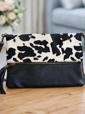 Svart handväska med koskinnsmönster - Snygg handväska i äkta läder med svart botten och lock i svartvitt koskinnsmönster. Väskan har guldfärgad dragkedja och dekorativ tofs. Tillverkad i Italien och har justerbar axelrem. Perfekt för dig som gillar djurmönstrat och trendiga detaljer.